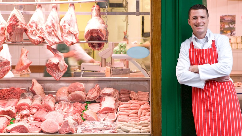 Butcher in red apron standing outside butcher shop with meat seen through window