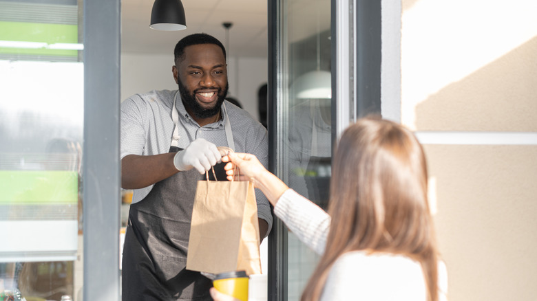 Smiling man hands brown takeout bag to customer through window.