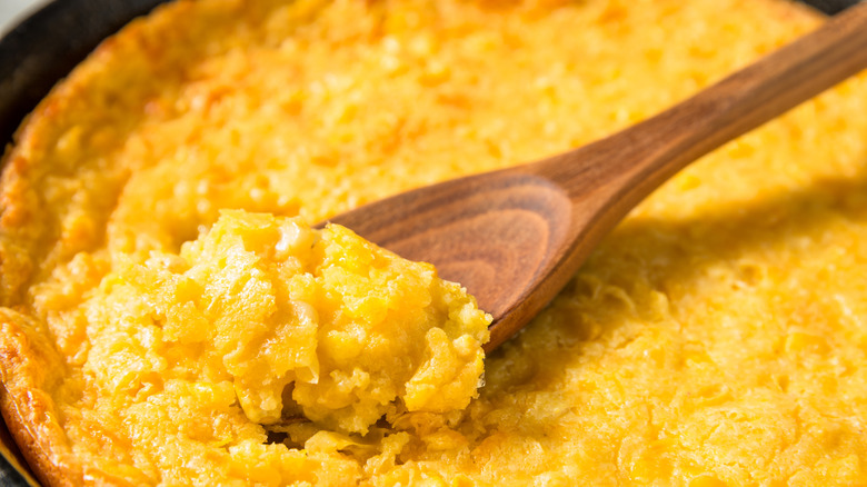 A close up for corn pudding being spooned out of a cast iron skillet with a wooden spoon