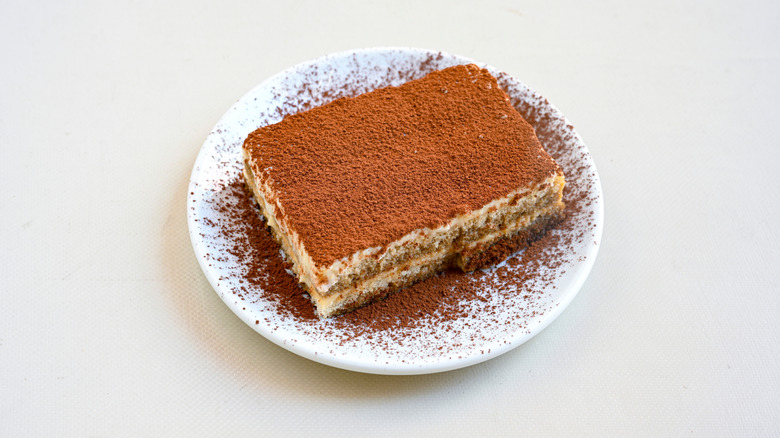 A square of tiramisu on top of a white plate, with lots of powdered cocoa on top