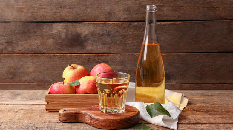 Glass of apple cider on a small wooden cutting board, with a bottle of apple cider and a box of apples behind it, all on a wooden background