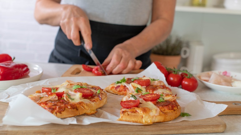 a cooked pizza on parchment pape on a wooden cutting board, a woman slices tomatoes in the background