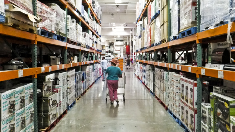 A shopper pushes cart though Costco aisle with many stacked goods.
