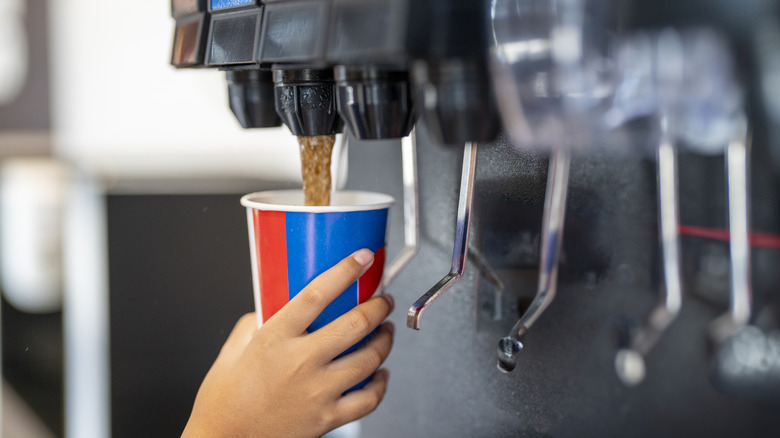 A hand fills a paper cup at a soda fountain