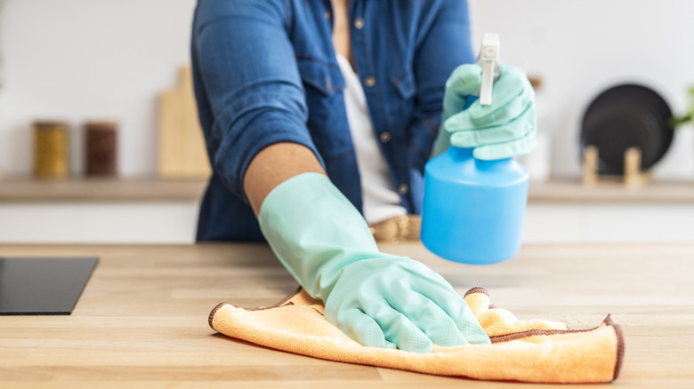 A close-up of someone wiping down a wooden tabletop with a cloth and spray bottle, wearing blue rubber gloves