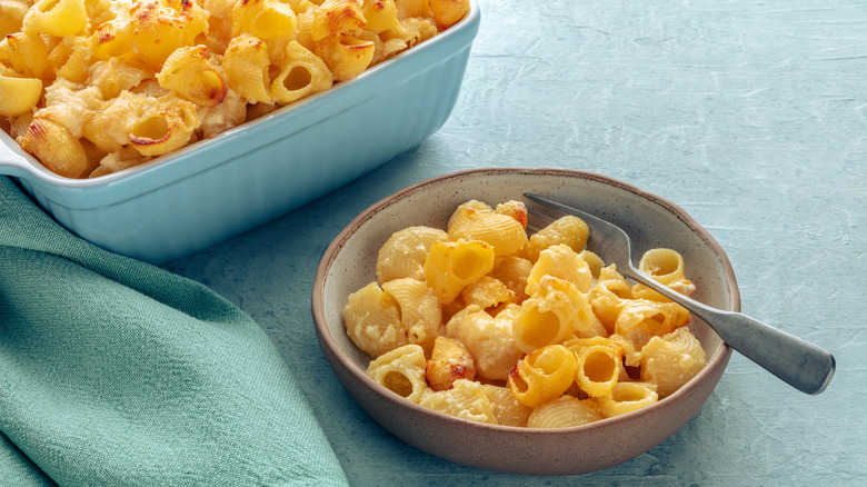 A close-up of a small bowl of mac and cheese with a fork, and a casserole dish full of mac and cheese in the background