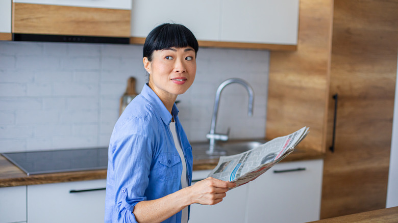 Woman stands in kitchen while holding newspaper.