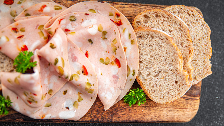 Sliced olive loaf deli meat next to a few pieces of bread on a wooden cutting board