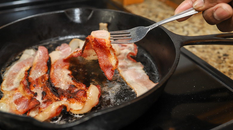 Bacon cooking in a pan with someone holding up one strip with a fork