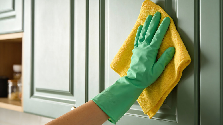 Hand in green glove cleaning a paneled kitchen cabinet with a yellow cloth