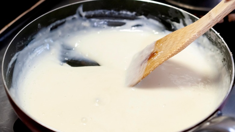 Person stirring béchamel sauce in black pan with wooden spoon