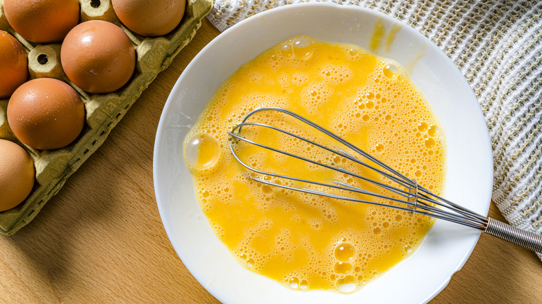Raw scrambled eggs and metal whisk in white plate with unbroken eggs in a carton in background