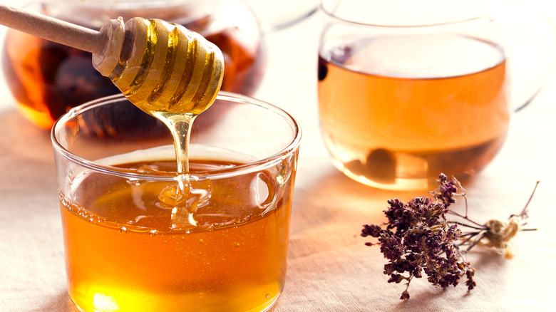 Honey drizzling from a wooden honey want into a glass of honey, with a second glass of honey and some dried flowers to the side