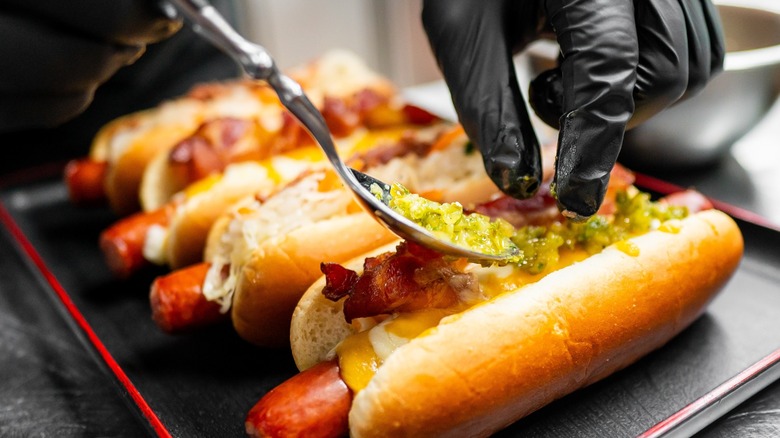 a gloved hand applies toppings to hot dogs