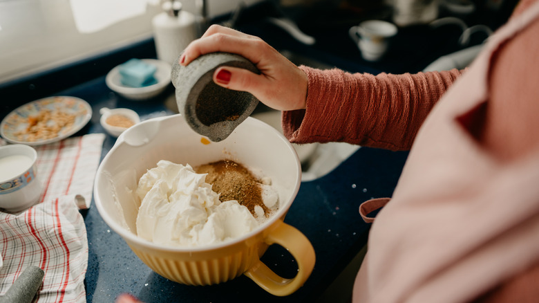 Person in apron pouring spices into a large yellow mixing bowl with other ingredients.
