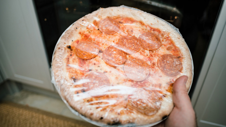 hand holds a plastic-wrapped frozen pizza