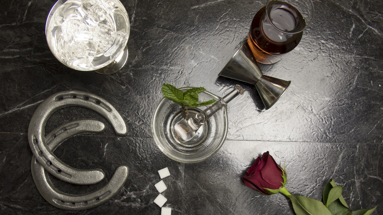 Mint julep glass with tumbler, horseshoes, bourbon, and a rose on dark background