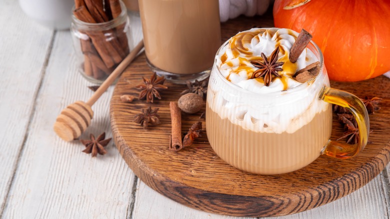 A close-up of a pumpkin spice latte in a glass mug sitting on a round wooden plate, with various spices like cinnamon and nutmeg scattered around