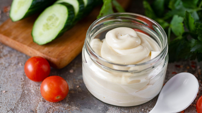 Mayonnaise in open jar, alongside tomatoes and sliced cucumber