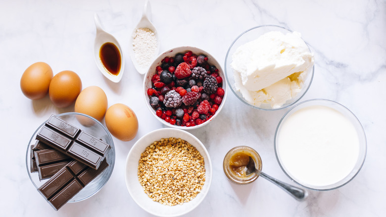 chocolate, eggs, berries, butter, milk and other cake ingredients laid out on a white marble countertop.
