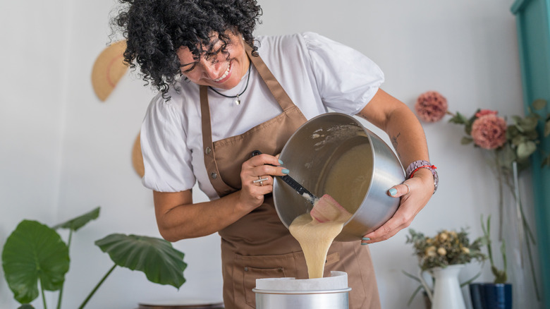 woman with curly black hair pouring cake batter into a bowl and smiling with flowers and plants behind her
