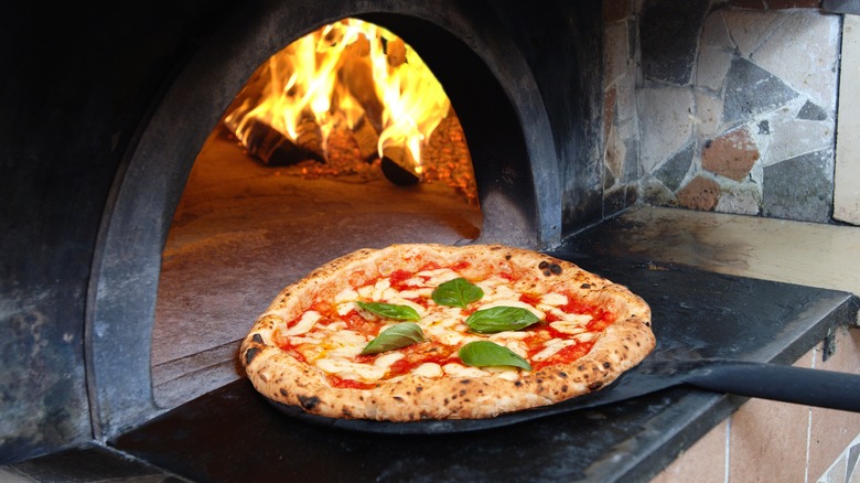 A margherita pizza being taken out of a wood-burning pizza oven with a black pizza peel