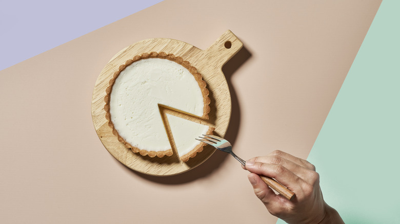 Overhead shot of cheesecake on wooden board, hand poised with fork above.