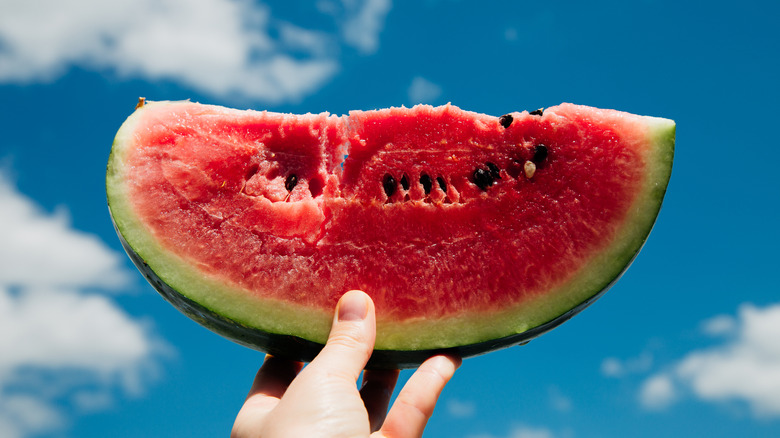 a hand holds watermelon aloft against the sky
