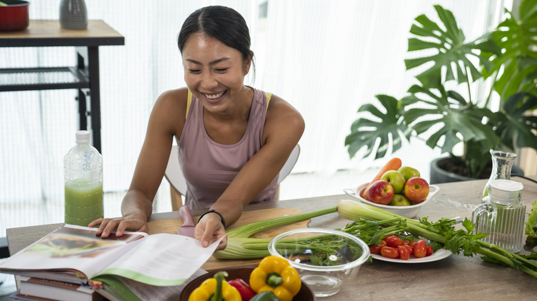 Someone reading a cookbook in a kitchen, with fresh produce like celery and bell peppers on their table