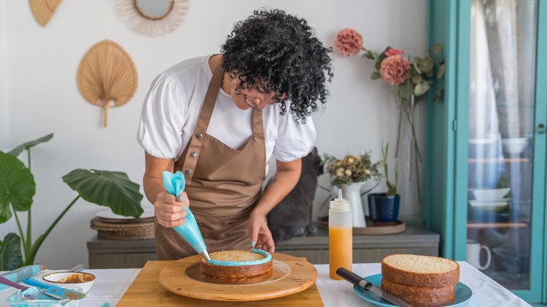 A woman frosting a cake layer with two more layers on a plate to the side