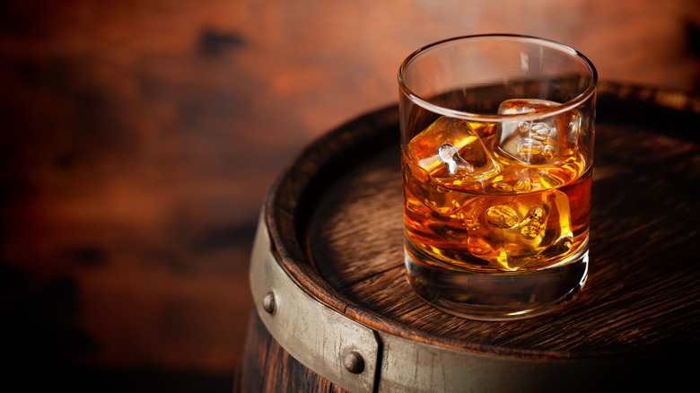 Glass of whiskey with ice cubes on wood barrel.
