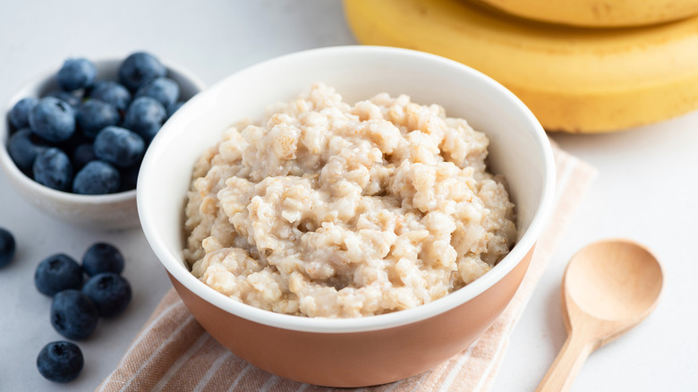 A bowl of oatmeal sitting on a wooden cutting board, next to a smaller bowl of blueberries, some bananas, and a wooden spoon
