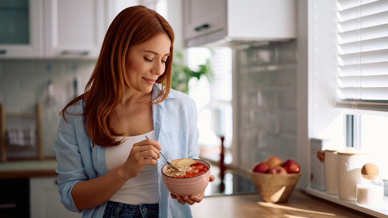 A woman standing in her kitchen eating oatmeal with fruit