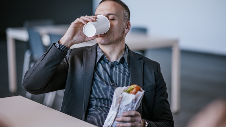 a professional drinks coffee and eats a sub sandwich