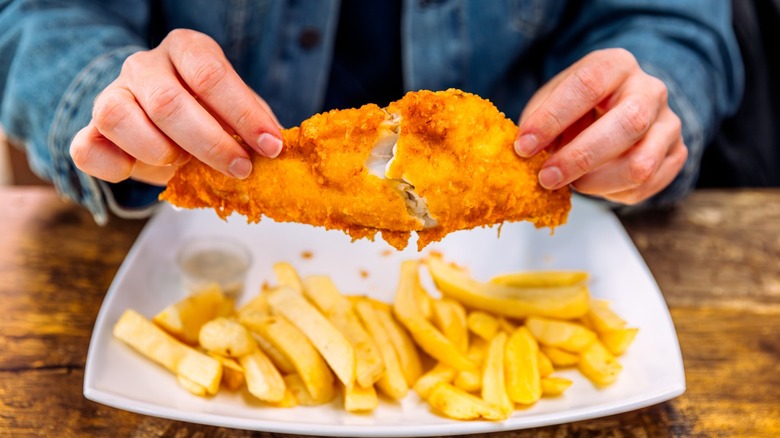 Hands hold a fish filet over a plate of french fries