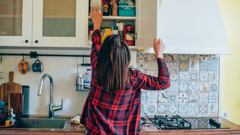 Person reaching up into cluttered kitchen cabinet, seen from behind.