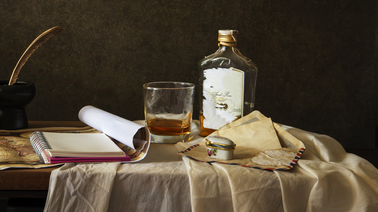 Still life with whiskey bottle, glass, notebook, and quill pen.