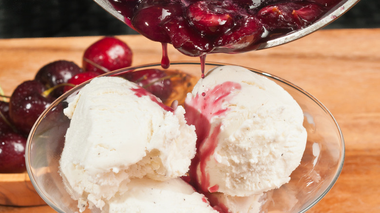 a bowl of ice cream with cherries in syrup being poured on top and cherries in the background, on a wooden table