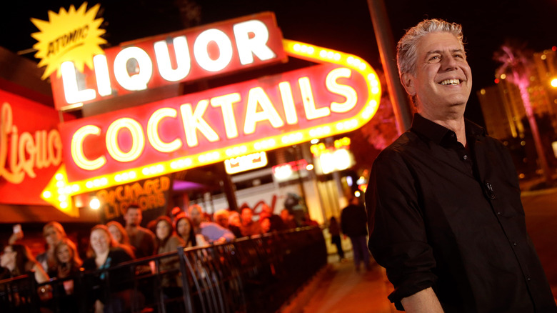 Anthony Bourdain standing in front of a neon sign that reads 'atomic liquor cocktails'