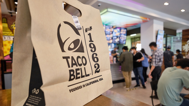 Taco Bell paper bag closeup with blurred customers in background.