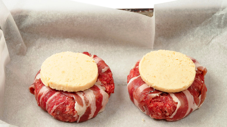 Two bacon-wrapped burger patties sit on parchment, a round of butter atop each