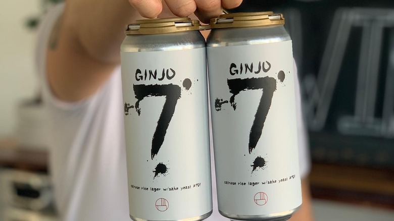person holding up two beer cans with a white label that reads 'ginjo 7 calrose rice lager w/sake yeast #701'