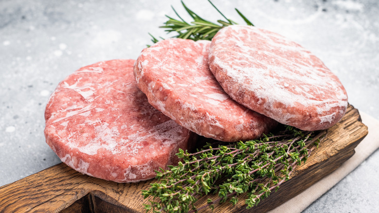 Three frozen hamburger patties on a wooden chopping board