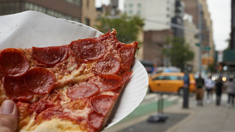 Person carrying thin pizza slice on paper plate in the city