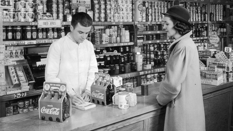 A shopper waits as grocery clerk writes up her bill, two six-packs of Coca-Cola next to her, 1935.