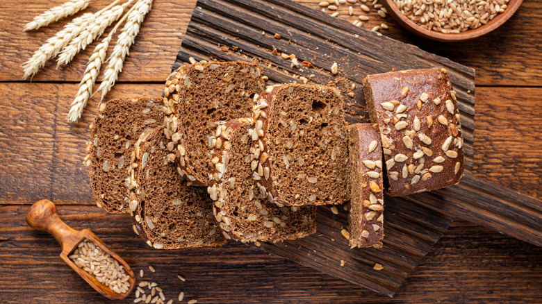 Sliced whole grain rye bread on a wooden cutting board, with wheat stalks and other grains scattered around