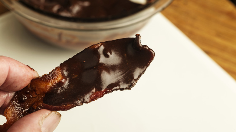 A close-up of a strip of bacon dipped in chocolate, with a bowl of chocolate in the background