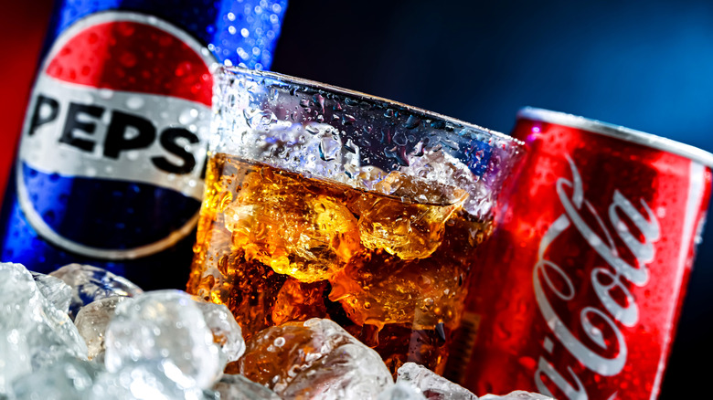 Cans of Pepsi and Coke with full glass and ice in foreground.