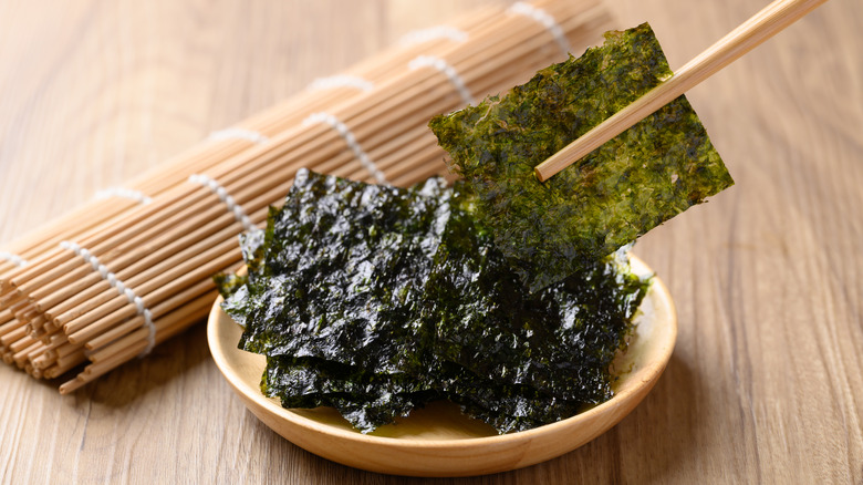 A close-up of someone holding a seaweed snack with chopsticks, and a small wooden bowl filled with more seaweed snacks