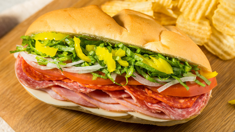 A hoagie with italian meat, tomatoes, onions, lettuce, and pickled peppers is on a cutting board.